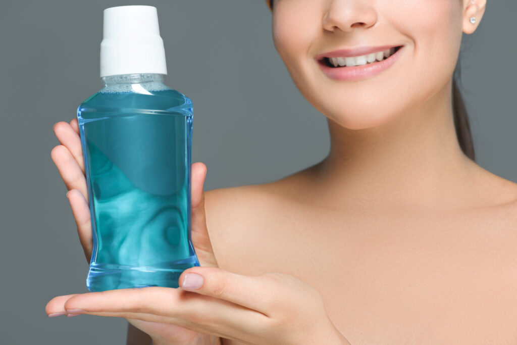 partial view of smiling woman with bare shoulders holding mouthwash isolated on grey