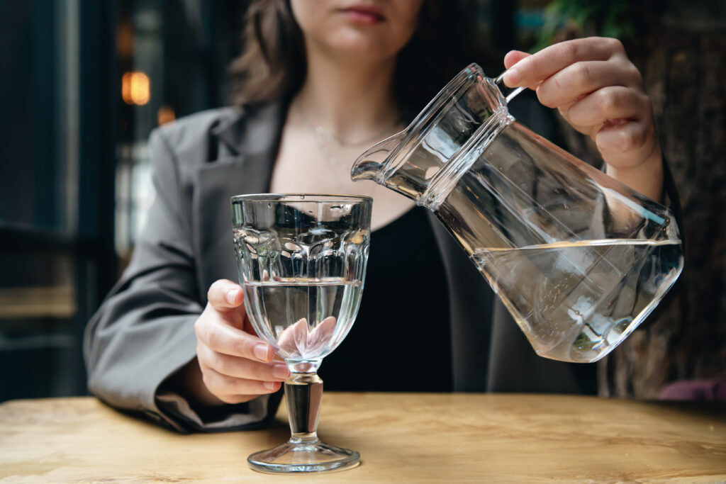 Close-up, a woman pours water from a glass jug into a glass goblet in a cafe.